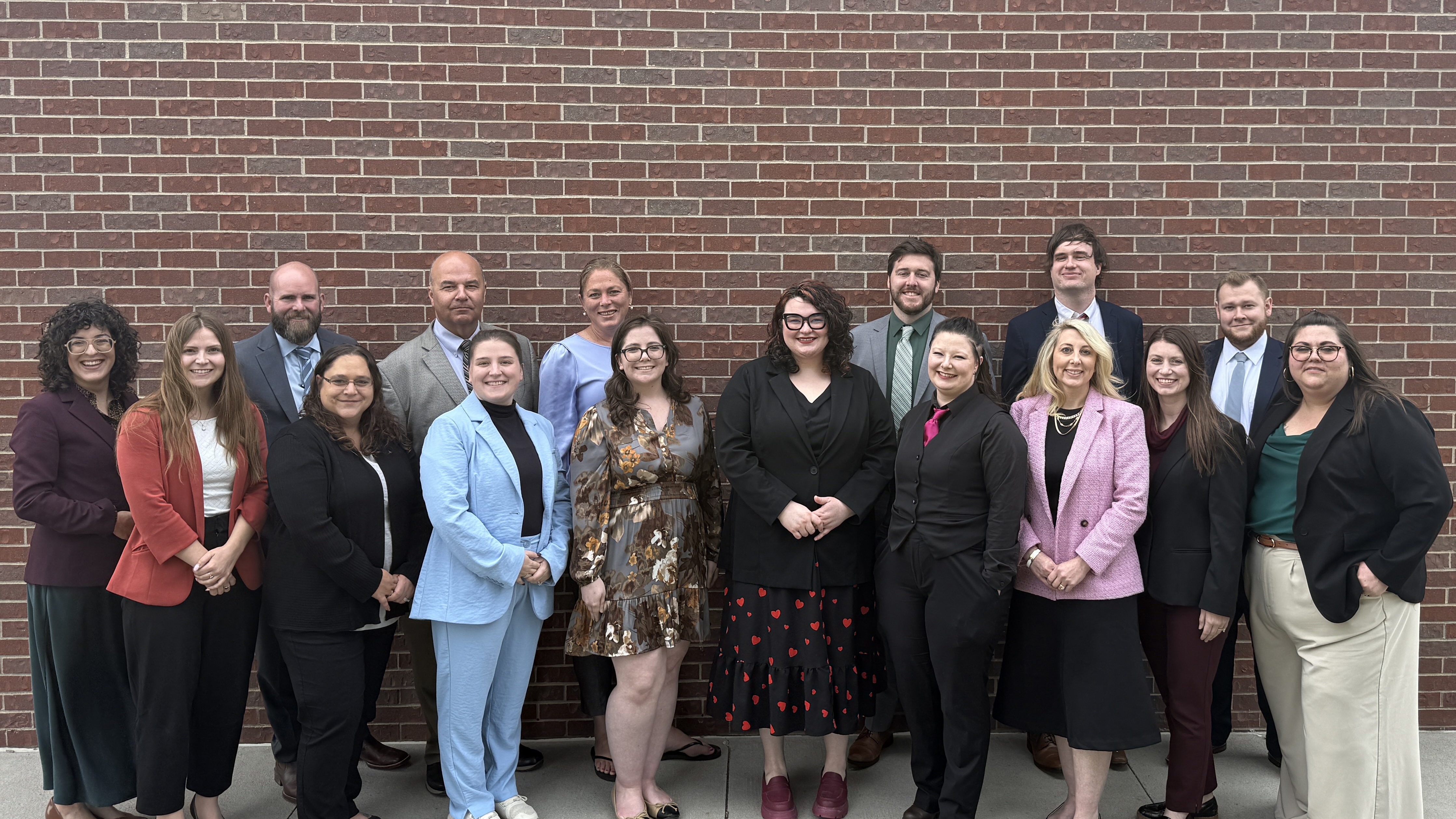 Sixteen attorneys in formal attire pose for a group photo in front of a brick wall.