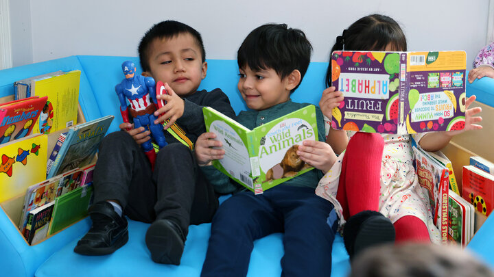 Three young children sit together, one holding a Captain America doll and two holding books