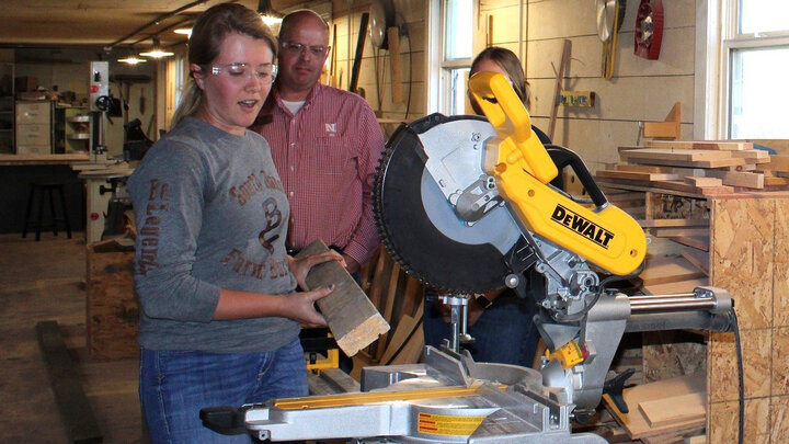 A young woman wearing safety goggles holds a board in front of a saw as a man and woman watch.