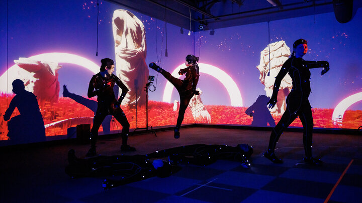 Three people move around in motion-capture suits, with two more on the ground, in front of an otherworldly background.