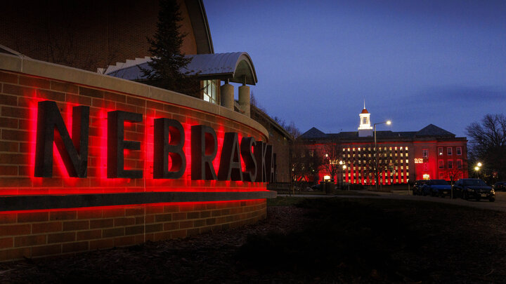 The word "Nebraska" glows in red on the sign east of the Van Brunt Visitors Center on the University of Nebraska–Lincoln's City Campus. Love Library South glows red in the background.