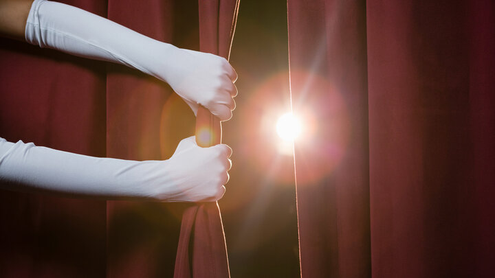 Two hands wearing long white gloves pull back a theater curtain as a spotlight shines through.