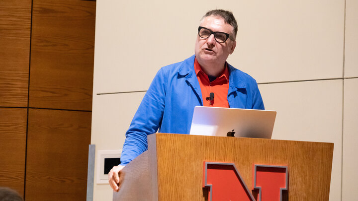 Jeffrey L. Day, professor of architecture at the University of Nebraska–Lincoln, speaks at a lectern in the Nebraska Union's Swanson Auditorium. He is wearing a longsleeve blue shirt over a red shirt.