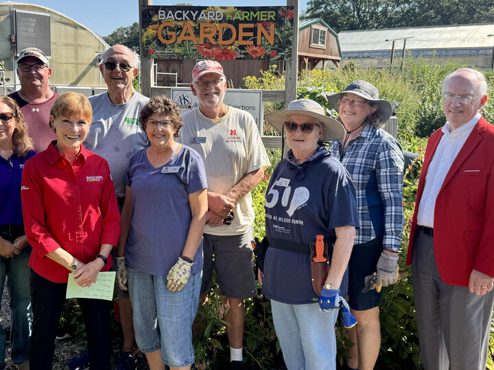 Nine people, seven in gardening clothes and two in business dress, pose for a photo in front of a garden.
